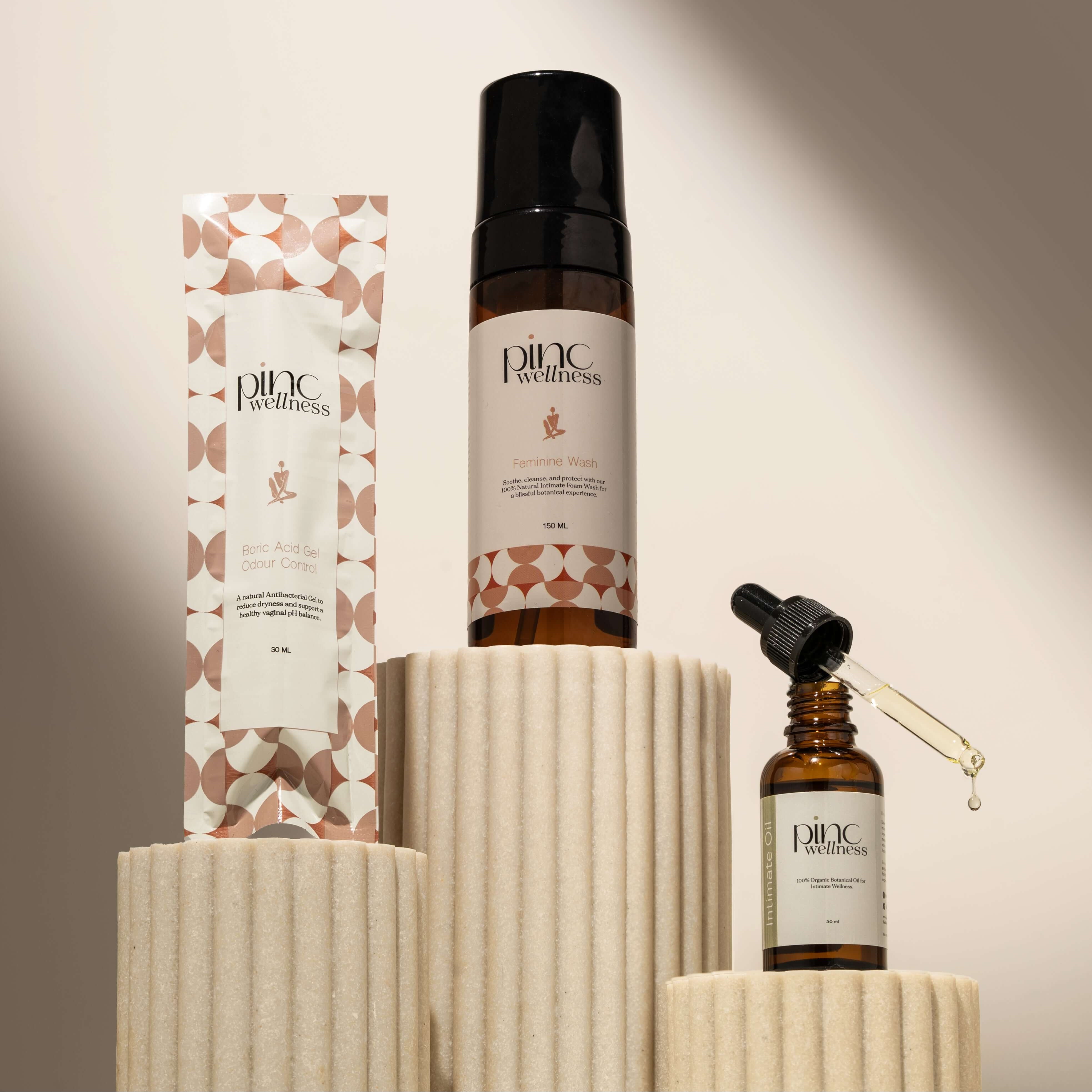 Three Pinc Wellness products on cream pedestals: wash, serum, gel.