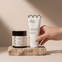 A hand holds a white tube of Pinac Wellness Intimate Lubricant against a warm beige background, next to a small jar of Pinac Wellness Lubricant Creamers resting on a textured stone block.