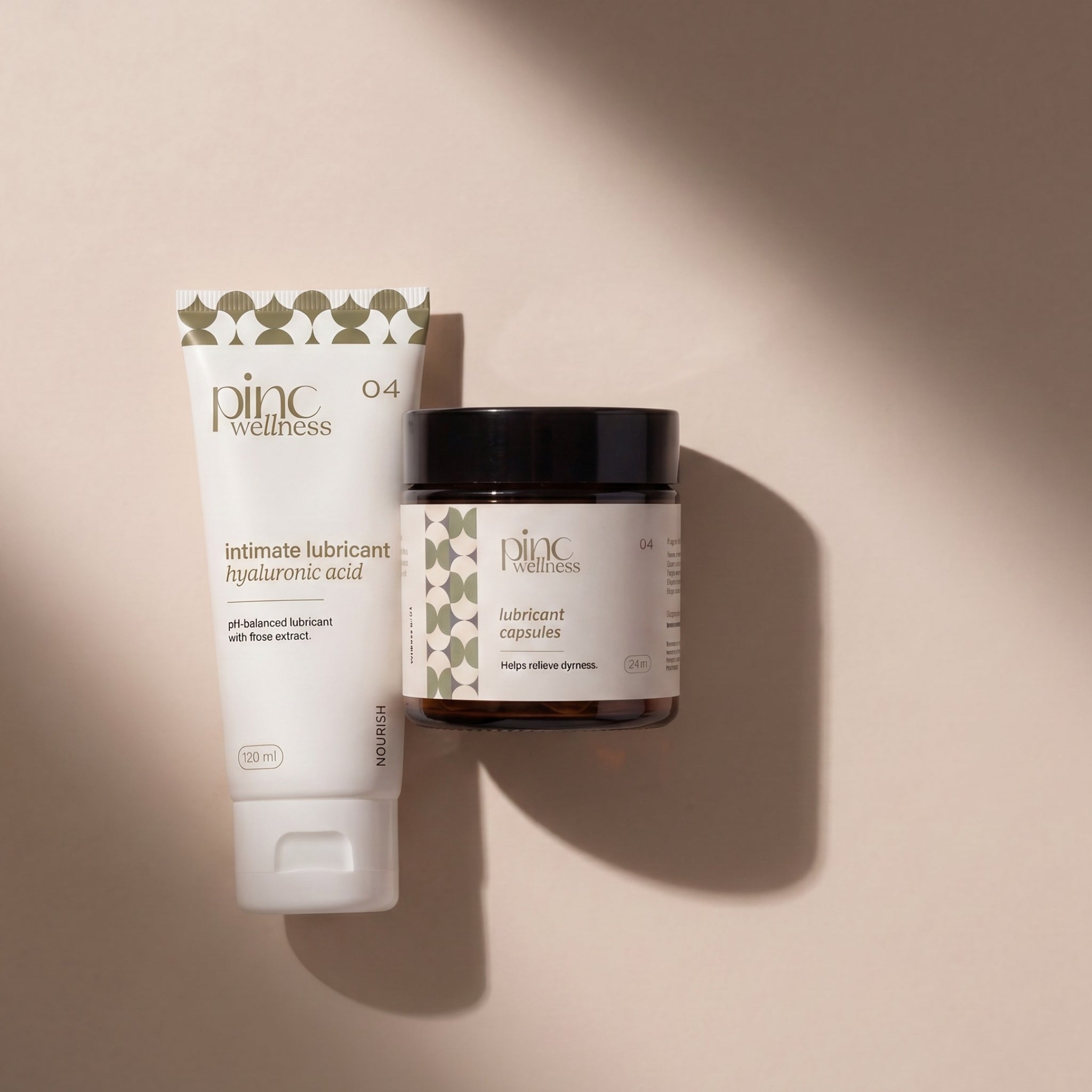 A white tube and a dark amber jar of Pinc Wellness lubricant products with a black lid, displayed on a warm beige background with soft diagonal shadows.